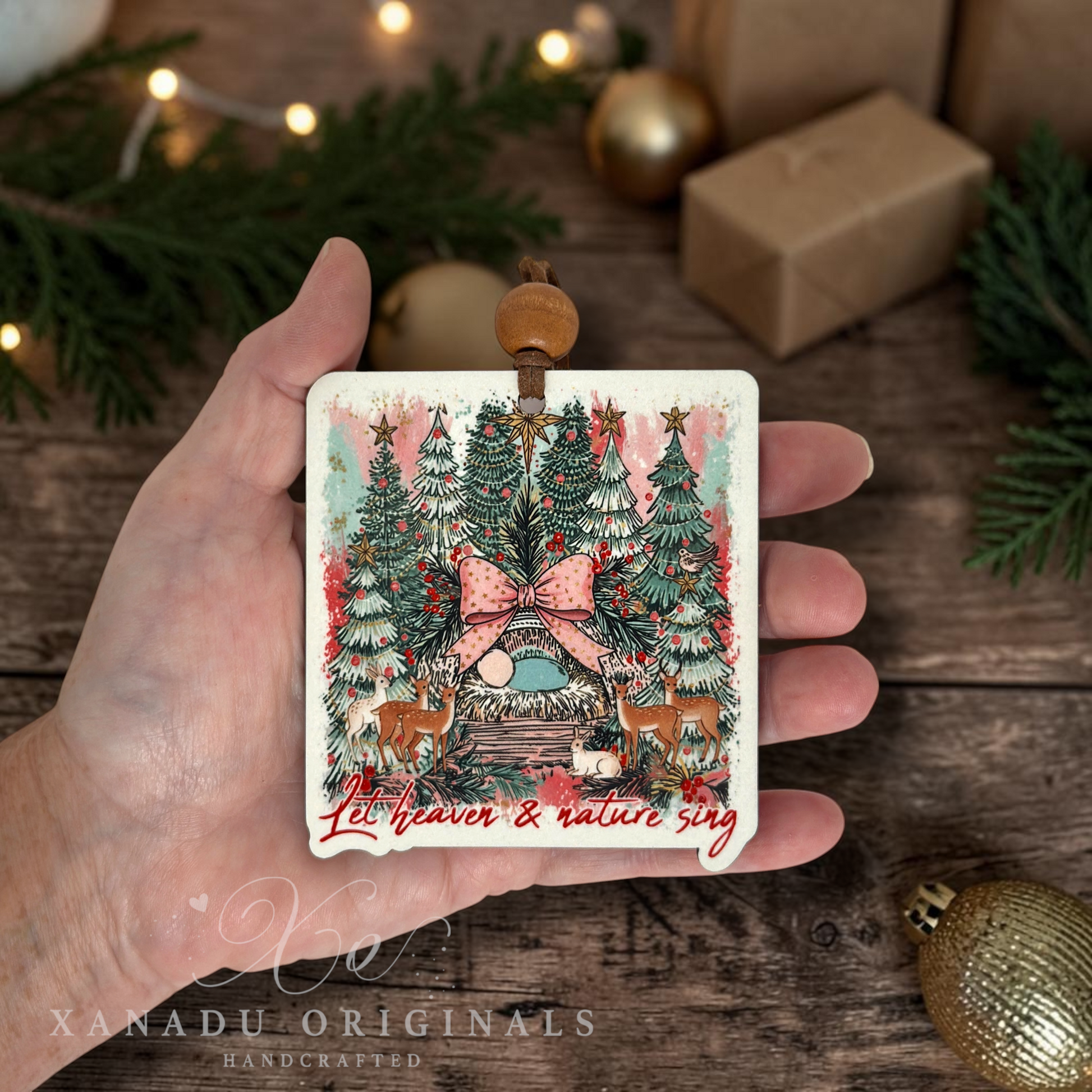 A hand holds a square Christmas ornament featuring an illustrated winter tree scene with a pink bow, deer, and the text 'Let heaven & nature sing.' The background shows a rustic wooden surface with holiday decorations, wrapped gifts, and string lights.