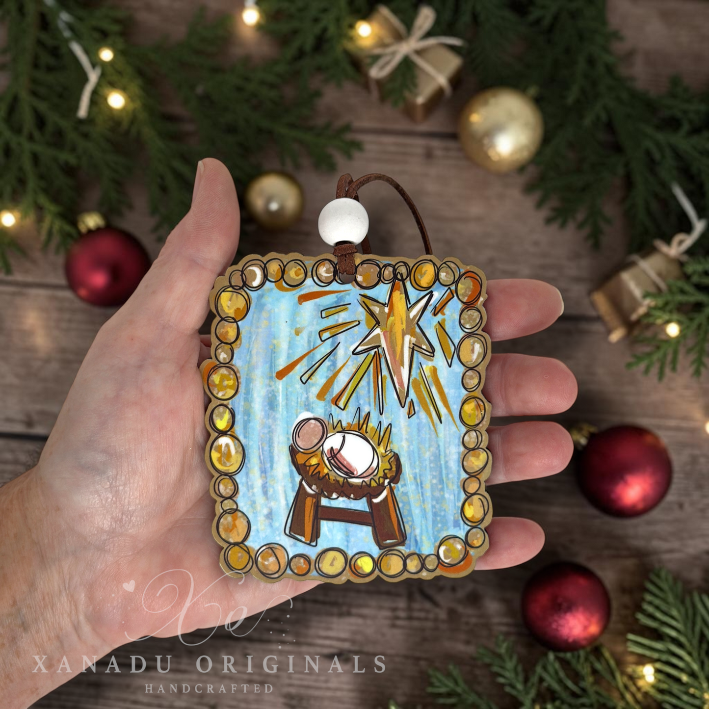 A hand holds a square Christmas ornament featuring a nativity scene with a blue background, gold starburst, and gold-toned beaded border. The scene is styled with holiday greenery, lights, and red ornaments on a wooden surface.