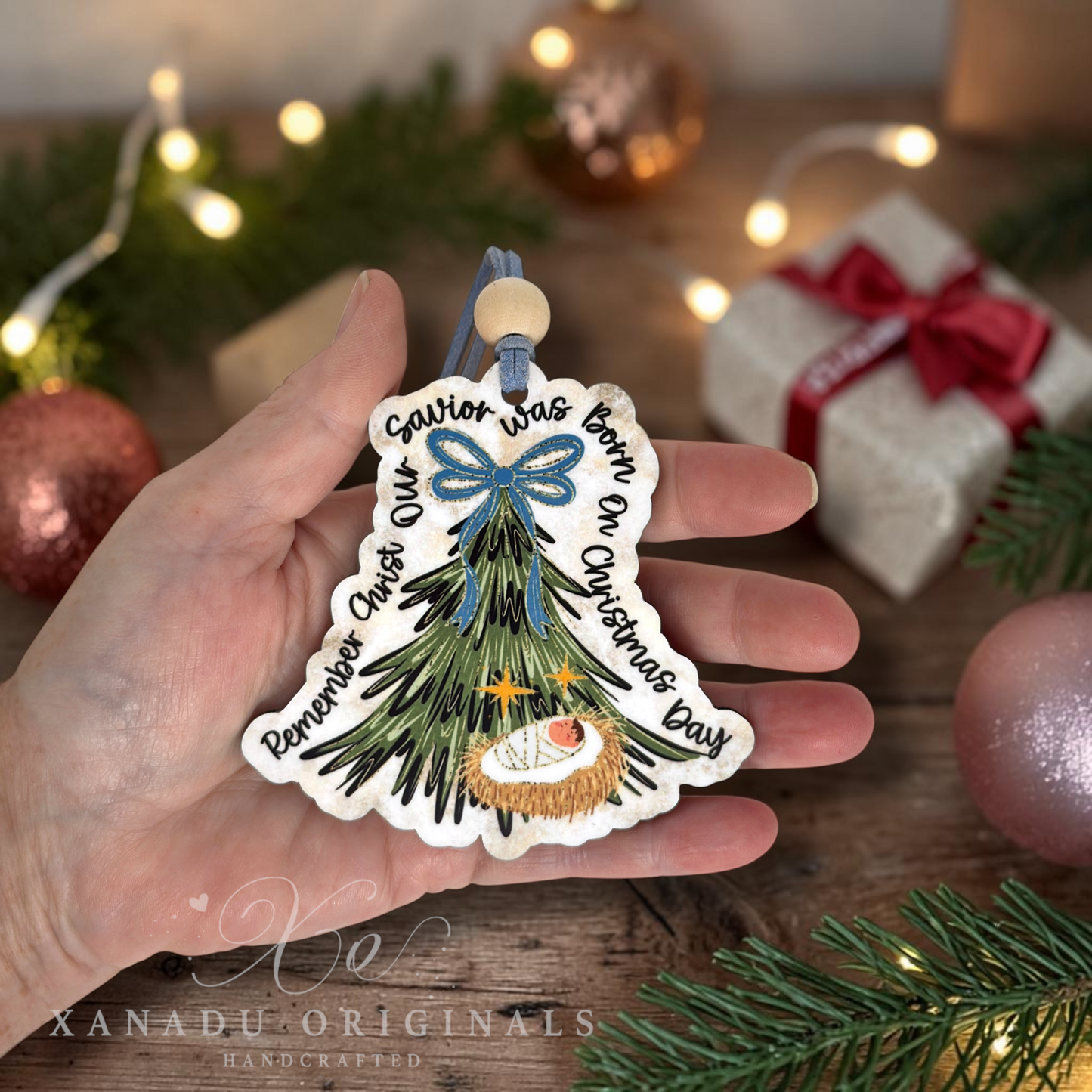 A hand holds a handcrafted Christmas ornament shaped like a Christmas tree with a white border and printed text reading 'Savior Was Born On Christmas Day' and 'Remember Christ Was Born On Christmas Day.' The ornament has green branches, a blue bow, and a small baby figure.