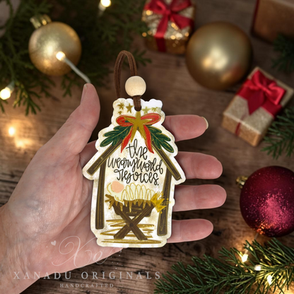 A hand holds a handcrafted Christmas ornament shaped like a stable, featuring the text 'The weary world rejoices' and a nativity scene. The background is a festive holiday setting with pine branches, gold ornaments, and wrapped gifts.