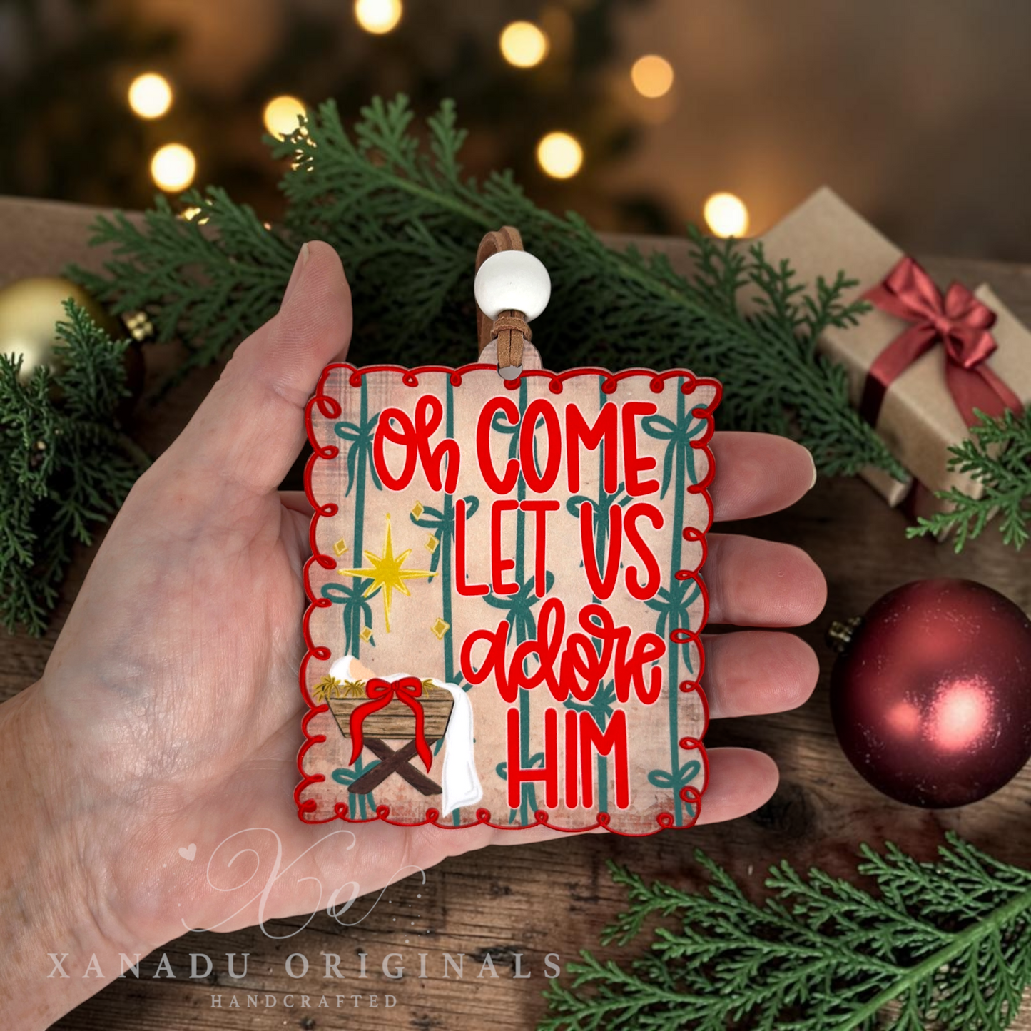 A hand holds a square Christmas ornament featuring the text 'Oh Come Let Us adore Him' with a manger scene and star. The background is a rustic holiday setting with pine branches, warm bokeh lights, a wrapped gift, and a red ornament.