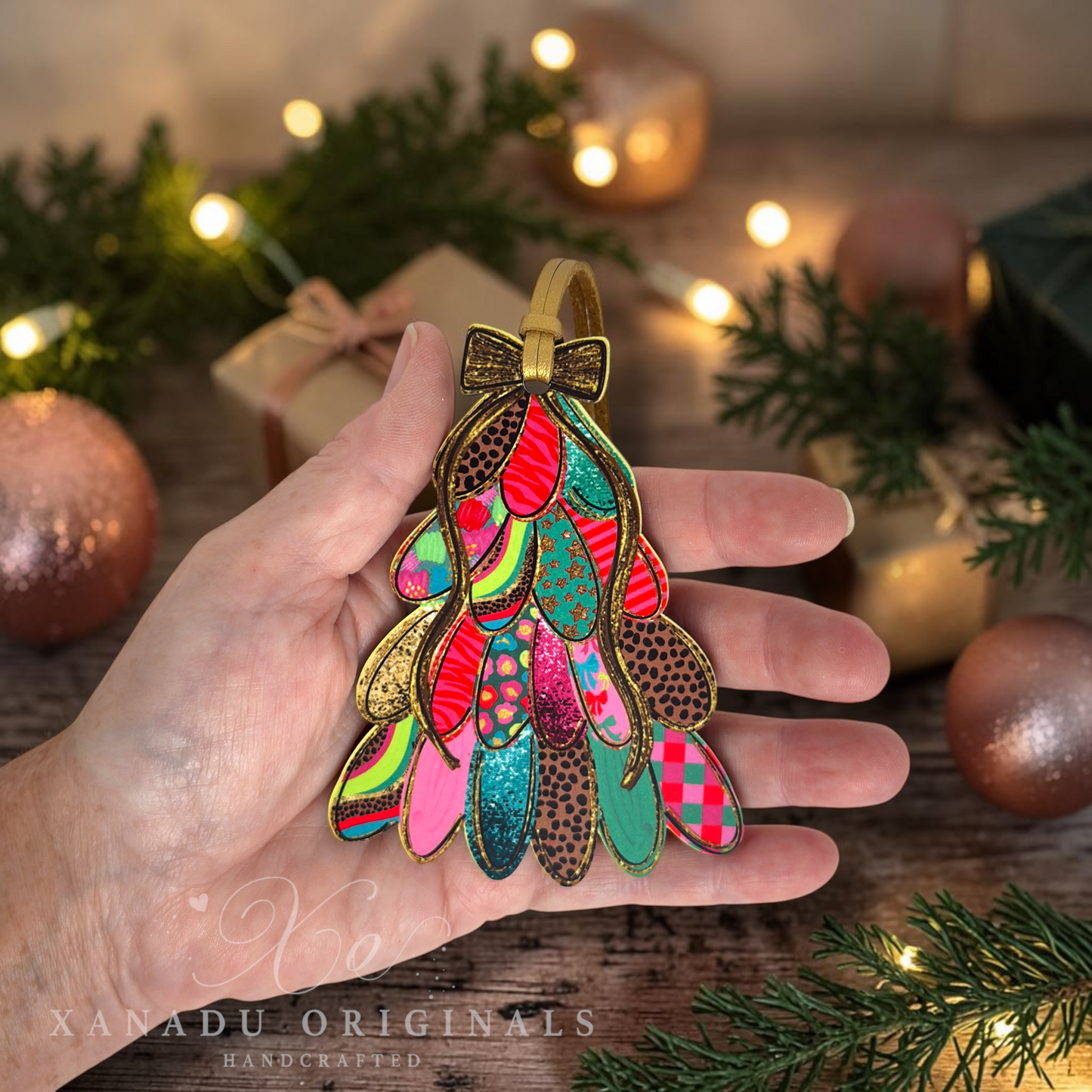 A hand holds a Christmas tree ornament made from colorful patterned paper pieces with gold-tone metal outlines, set against a warm, festive background with pine branches, lights, and ornaments. A watermark for 'XANADU ORIGINALS HANDCRAFTED' is visible in the lower left.