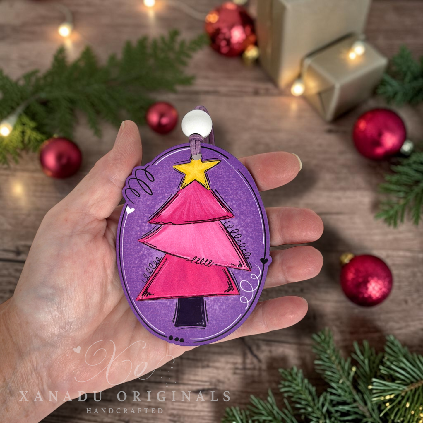 A hand holds a handcrafted oval Christmas ornament featuring a pink and magenta tree on a purple background with a yellow star, set against a rustic wooden table with holiday decorations.