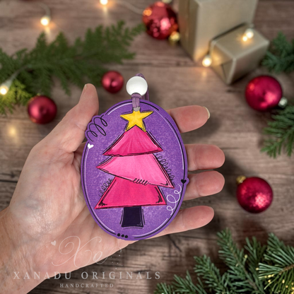 A hand holds a handcrafted oval Christmas ornament featuring a pink and magenta tree on a purple background with a yellow star, set against a rustic wooden table with holiday decorations.
