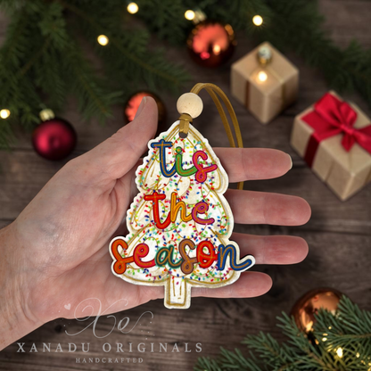 A hand holds a Christmas tree-shaped ornament with the phrase “tis the season” in colorful raised letters. The ornament has a white base with multicolored confetti and a gold hanging loop, photographed against a festive holiday background with pine branches, ornaments, and wrapped gifts.