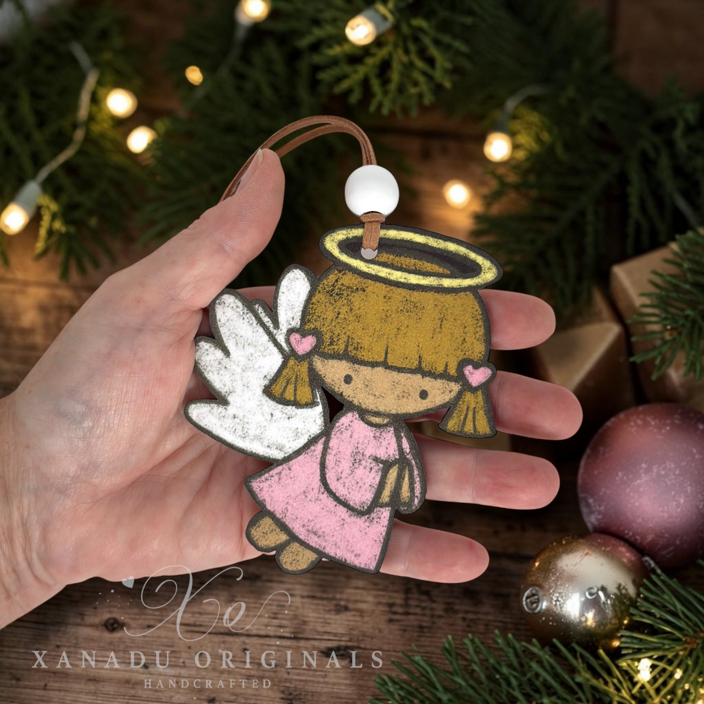 A hand holds a handcrafted angel ornament with a brown leather loop, pink dress, gold hair and halo, and white wings. The background features a wooden surface decorated with green pine branches, warm fairy lights, and Christmas ornaments, with the brand name 'XANADU ORIGINALS' visible in the bottom left corner.