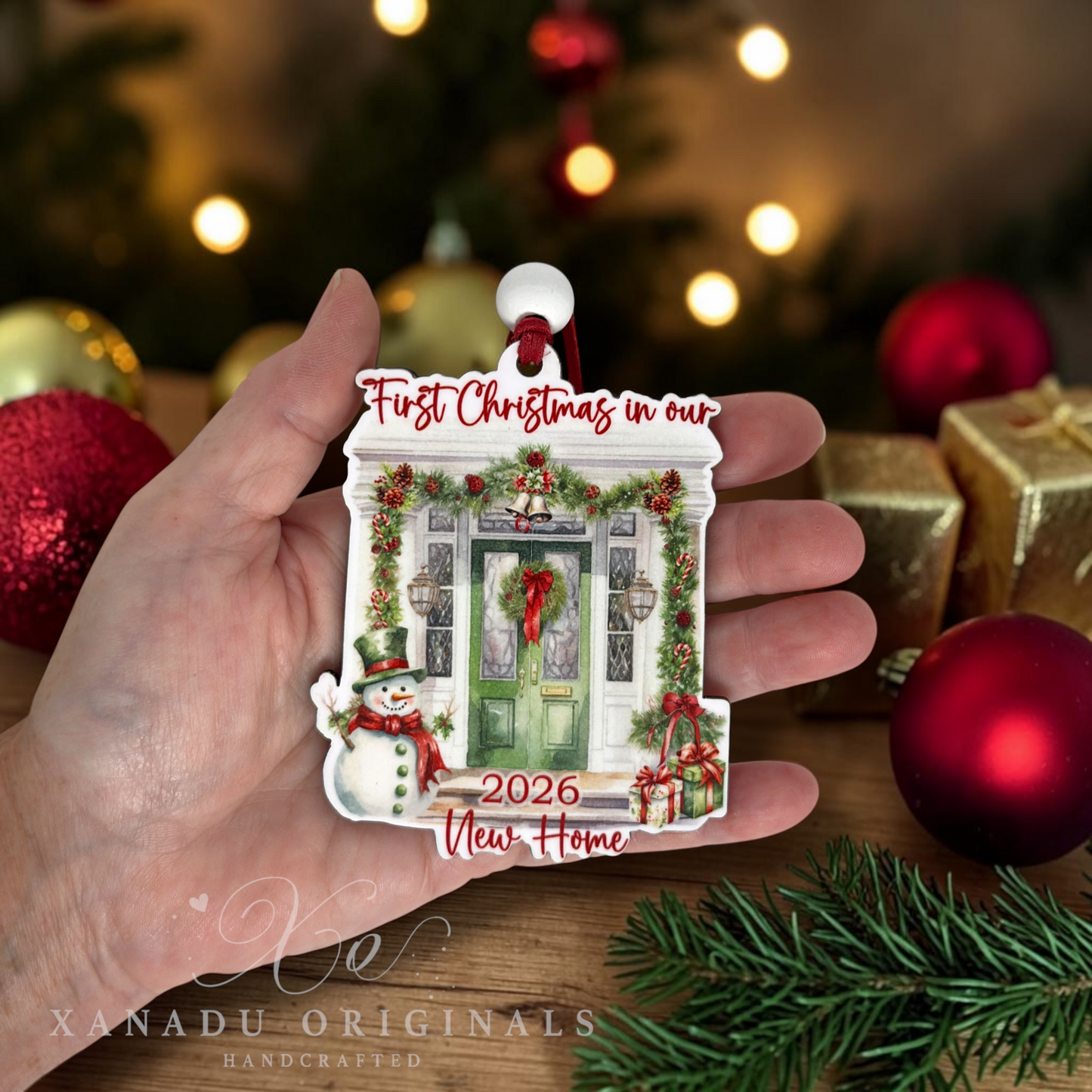 A hand holds a custom house-shaped Christmas ornament featuring a decorated green front door, a snowman, and the text 'First Christmas in our New Home 2026'. The background shows a warm, blurred Christmas tree and ornaments.