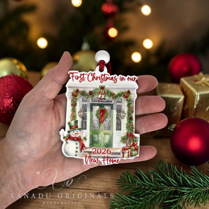 A hand holds a custom house-shaped Christmas ornament featuring a decorated green front door, a snowman, and the text 'First Christmas in our New Home 2026'. The background shows a warm, blurred Christmas tree and ornaments.