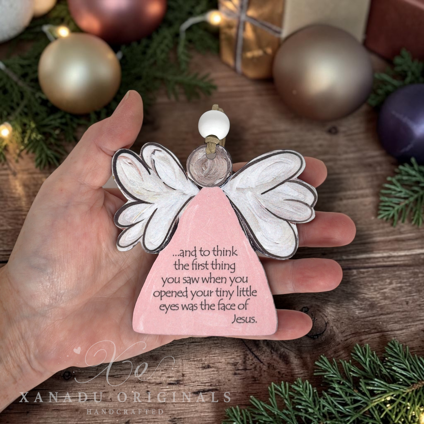 A hand holds a handcrafted Christmas angel ornament with a pink body, white wings, and printed memorial text, set against a rustic holiday background with ornaments and pine branches.