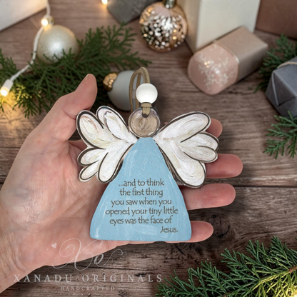 A hand holds a light blue angel-shaped ornament with white wings and a gold hanging loop, featuring engraved text that reads, and to think the first thing you saw when you opened your tiny little eyes was the face of Jesus'.