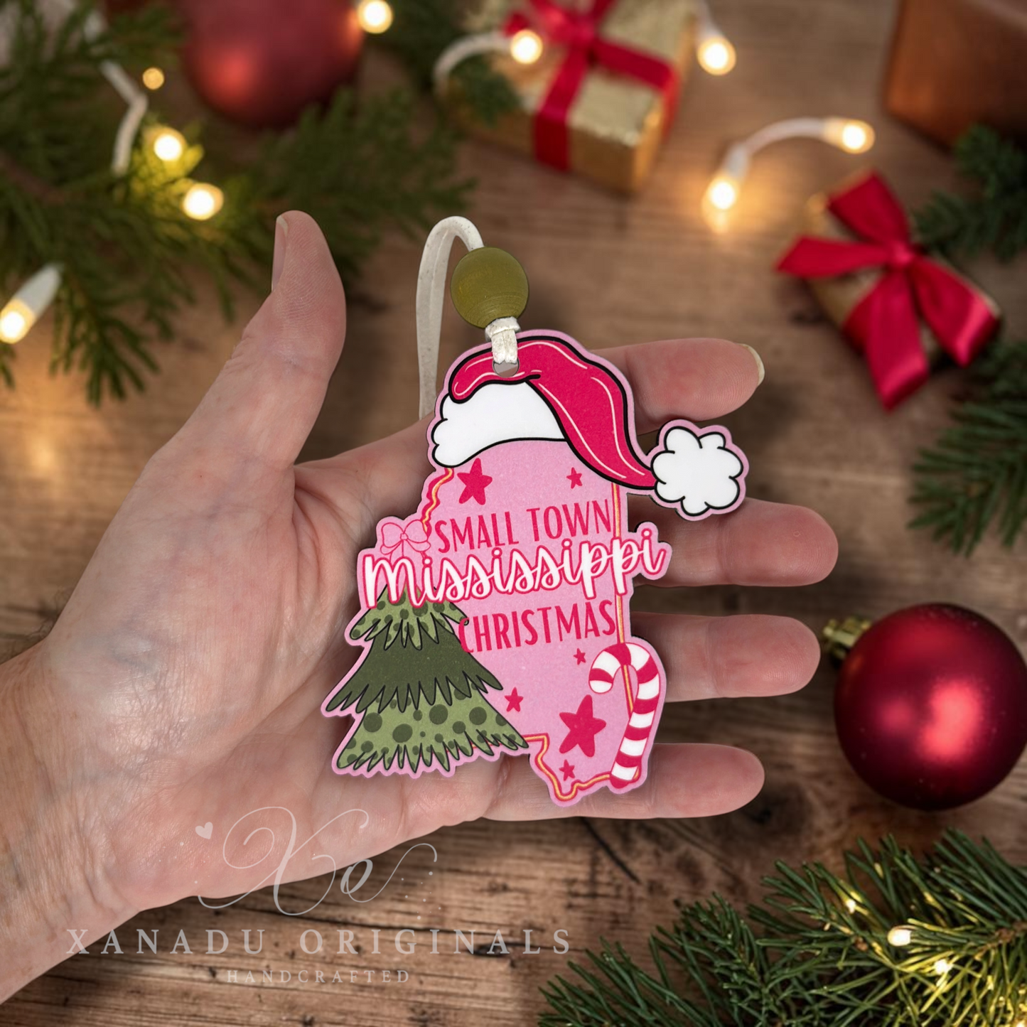 A hand holds a pink Christmas ornament shaped like the state of Mississippi, decorated with a red Santa hat, green tree, candy cane, and the words 'Small Town Mississippi Christmas.' The background shows a wooden surface with holiday lights, pine branches, and red ornaments.