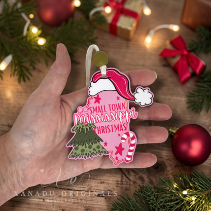 A hand holds a pink Christmas ornament shaped like the state of Mississippi, decorated with a red Santa hat, green tree, candy cane, and the words 'Small Town Mississippi Christmas.' The background shows a wooden surface with holiday lights, pine branches, and red ornaments.