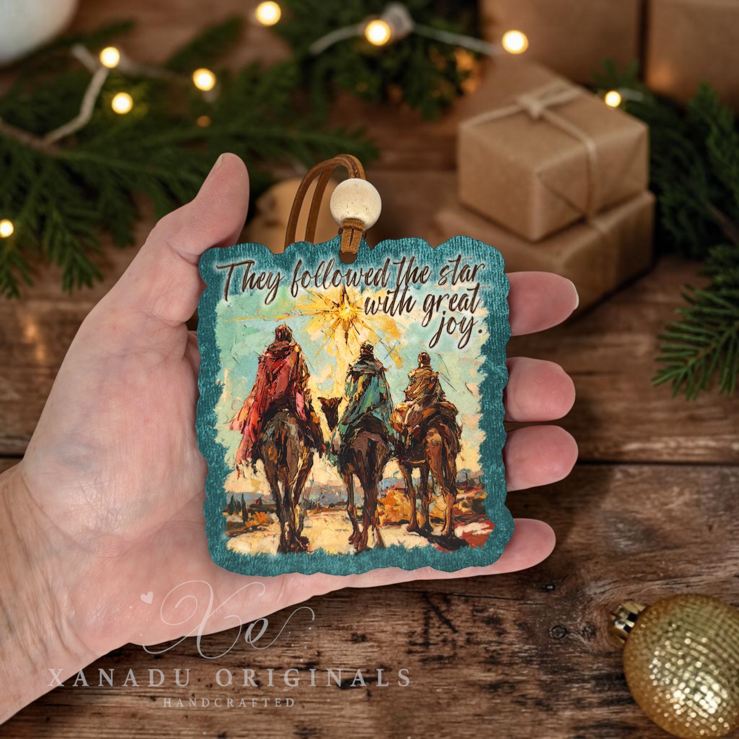 A hand holds a square decorative ornament featuring an illustrated scene of three camels and riders under a star, with the text 'They followed the star with great joy.' The background shows a rustic wooden surface with holiday greenery, string lights, wrapped gifts, and a gold ornament.