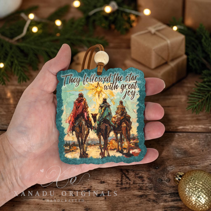 A hand holds a square decorative ornament featuring an illustrated scene of three camels and riders under a star, with the text 'They followed the star with great joy.' The background shows a rustic wooden surface with holiday greenery, string lights, wrapped gifts, and a gold ornament.