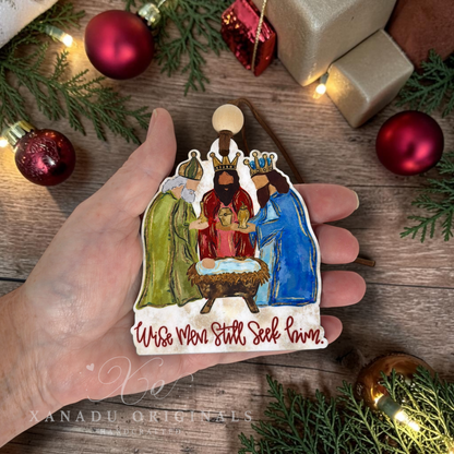 A hand holds a hand-painted Christmas ornament depicting the Holy Family in a nativity scene, with the text 'Wise Men Still Seek Love.' The background is a rustic wooden surface decorated with red ornaments, pine branches, and wrapped gifts.