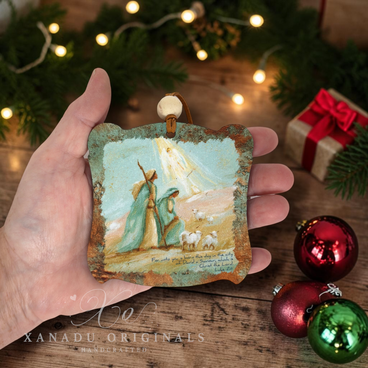 A hand holds a rustic Christmas ornament featuring a painted scene of the Three Wise Men and a sheep, with a warm glowing angel light in the background. The ornament has a distressed, weathered border and is shown against a cozy holiday backdrop with pine branches, fairy lights, a wrapped gift, and shiny red and green ornaments.