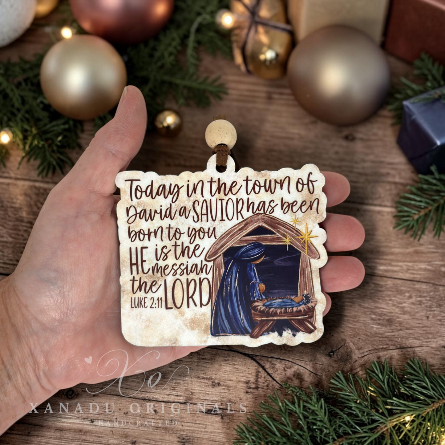 A hand holds a hand-painted wooden Christmas ornament featuring a nativity scene and the Bible verse 'Today in the town of David a Savior has been born to you, He is the Messiah the LORD Luke 2:11'. The background is a rustic wooden surface decorated with gold Christmas ornaments, pine branches, and wrapped gifts.