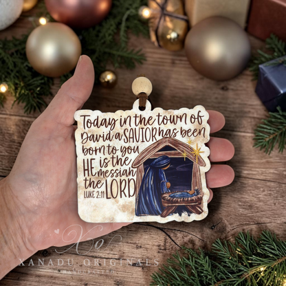 A hand holds a hand-painted wooden Christmas ornament featuring a nativity scene and the Bible verse 'Today in the town of David a Savior has been born to you, He is the Messiah the LORD Luke 2:11'. The background is a rustic wooden surface decorated with gold Christmas ornaments, pine branches, and wrapped gifts.