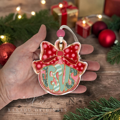 A hand holds a Christmas ornament shaped like a jingle bell, decorated with a red and pink polka-dot bow, gold trim, and pastel green and pink monogram lettering that says JESUS. The background shows a rustic wooden table with holiday decorations, including pine branches, red ornaments, and wrapped gift boxes.