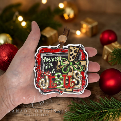 A hand holds a decorative Christmas ornament featuring the text 'The Real Gift Is Jesus' in stylized lettering with a red, green, and tan patterned background. The scene is decorated with red ornaments, pine branches, and warm holiday lights in the background.