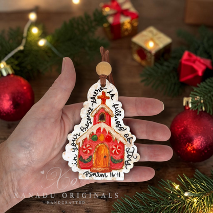 A hand holds a handcrafted Christmas ornament shaped like a church tree. The ornament is decorated with a red church, green leaves, candy canes, and black handwritten text.