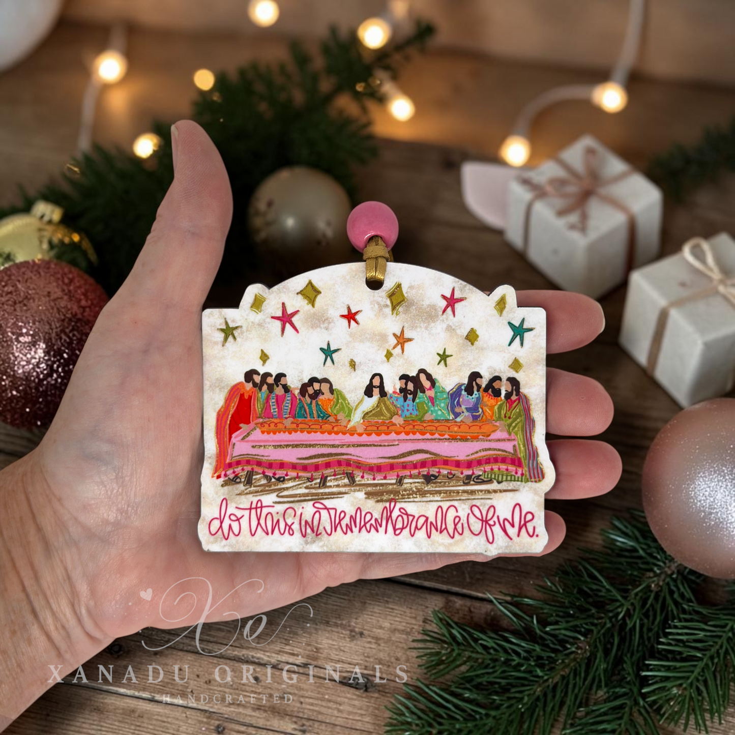 A hand holds a hand-painted Christmas ornament depicting the Last Supper with the text 'do this in memory of me' in red. The background is a cozy, festive tabletop setting with pine branches, warm string lights, and wrapped gift boxes.