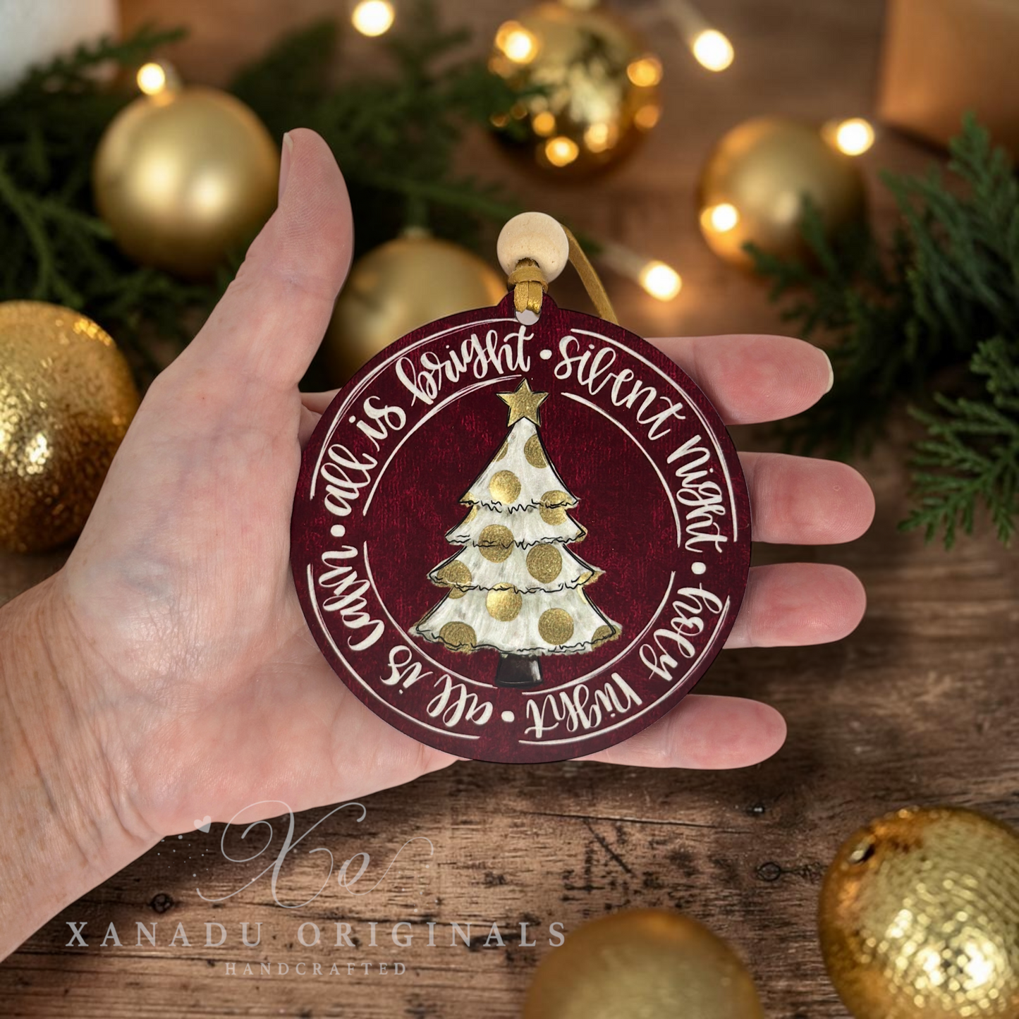 A hand holds a round maroon Christmas ornament featuring a white and gold decorated tree and the words 'all is bright - sweet night'. The background shows a rustic wooden surface with gold ornaments and green pine branches.