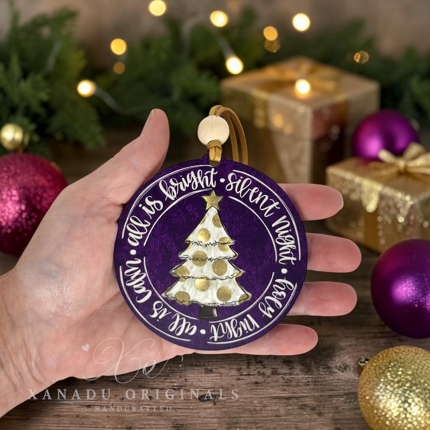 A hand holds a round purple Christmas ornament with a gold hanging loop. The ornament features a white and gold Christmas tree in the center and white script text around the edge reading 'all is quiet * night'.
