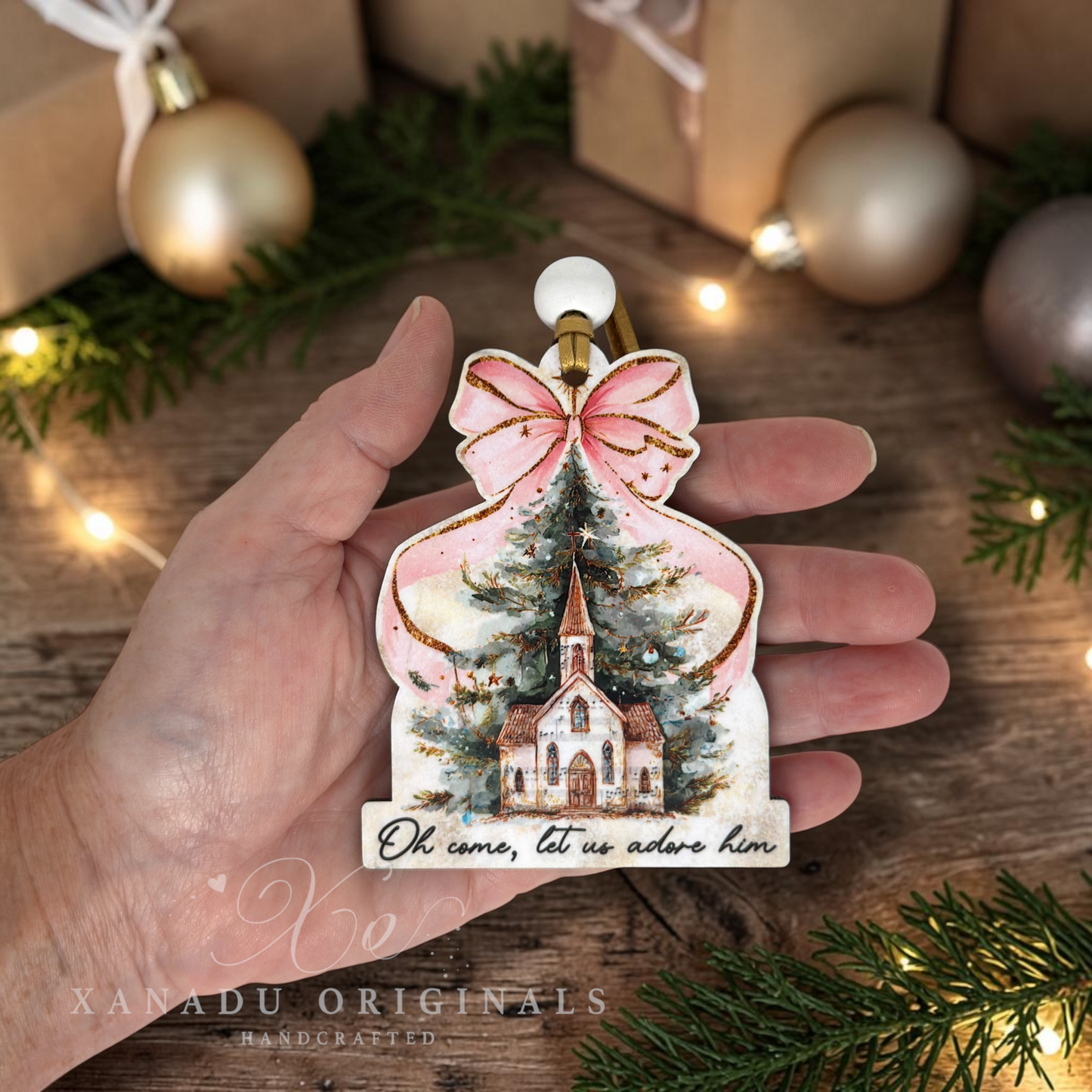 A hand holds a hand-painted Christmas ornament shaped like a church and tree, decorated with a pink bow and gold glitter accents, set against a rustic holiday background with wrapped gifts and lights.
