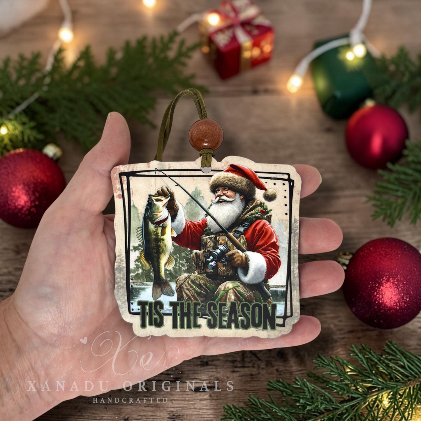 A hand holds a square Christmas ornament featuring a Santa Claus fishing and holding a fish, with the text 'TIS THE SEASON'. The background is a rustic wooden surface decorated with pine branches, red ornaments, and warm string lights.
