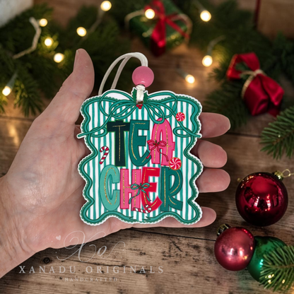 A hand holds a decorative Christmas ornament shaped like a gift box with the word "TEACHER" in layered pink, green, and black letters. The ornament has a white and green striped center, teal border, candy cane details, and a pink bead at the top.