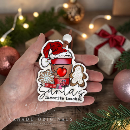 A hand holds a Christmas-themed die-cut ornament that reads 'Santa's favorite teacher,' decorated with a Santa hat, red drink cup, gingerbread man, snowflake, and gift motifs. The background shows a cozy holiday scene with wrapped gifts, lights, ornaments, and pine branches.