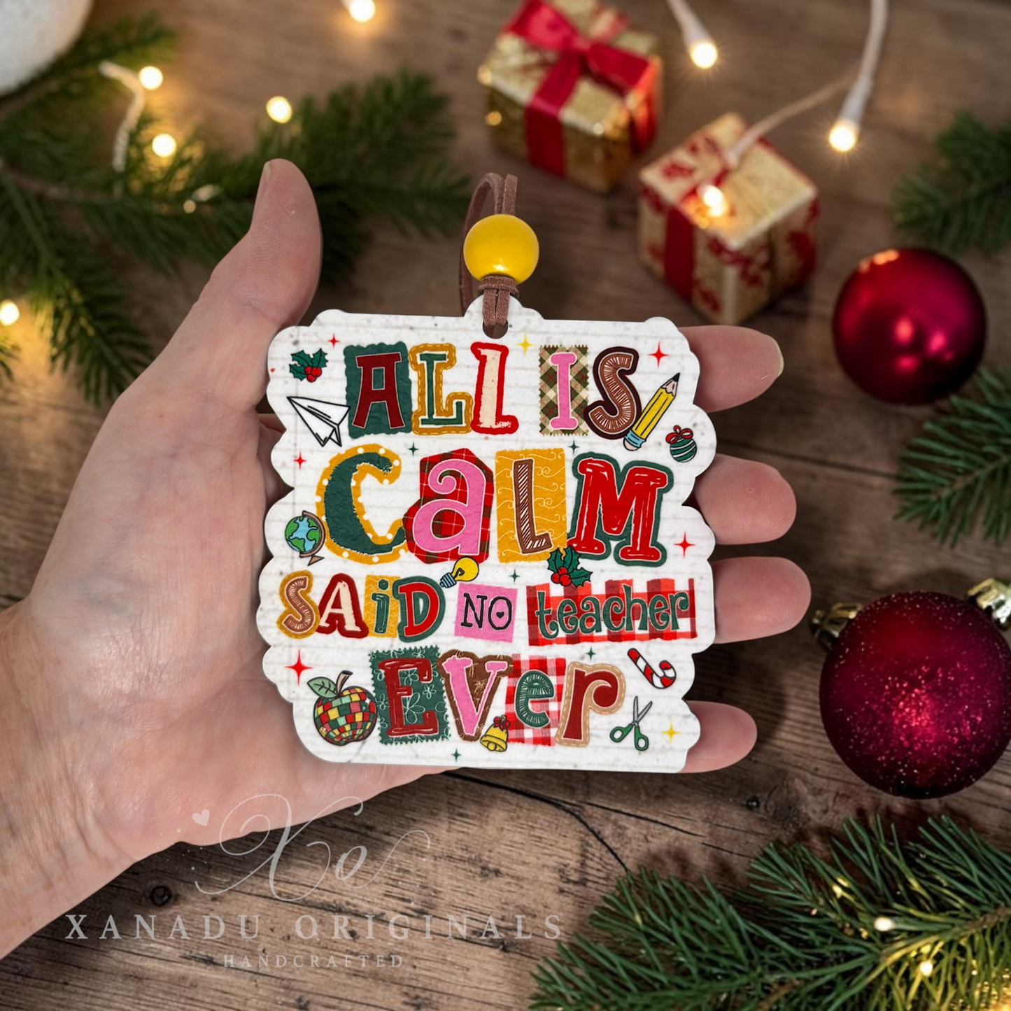 A hand holds a white scalloped ornament with colorful hand-lettered text reading 'ALL IS CALM SAID NO TEACHER EVER,' surrounded by Christmas greenery, ornaments, and wrapped gifts on a wooden surface.