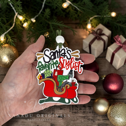 A hand holds a Christmas ornament shaped like Santa's sleigh with the text 'Santa's favorite Stylist' in festive lettering. The scene is decorated with pine branches, warm string lights, wrapped gift boxes, and red and gold ornaments on a rustic wooden surface.