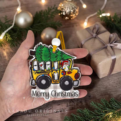 A hand holds a handcrafted Christmas ornament shaped like a yellow school bus carrying a green Christmas tree, with a Santa figure in the driver seat and the words 'Merry Christmas' at the bottom. The background shows a wooden table with holiday decorations, including pine branches, gold ornaments, and wrapped gifts.