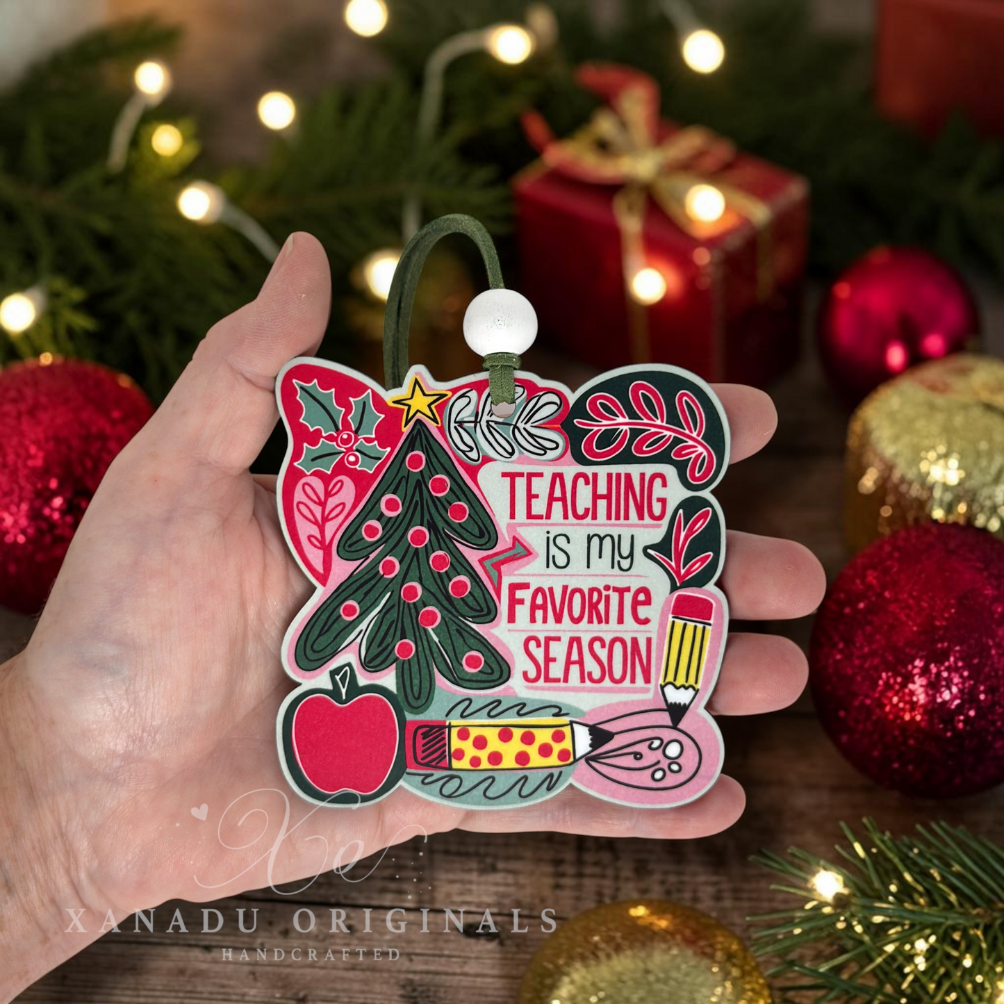 A hand holds a handcrafted Christmas ornament shaped like a chalkboard, featuring the text 'TEACHING is my FAVORITE SEASON' surrounded by festive illustrations of a Christmas tree, apple, pencil, and holiday motifs. The background is a cozy, blurred Christmas scene with lights, pine branches, and red ornaments.