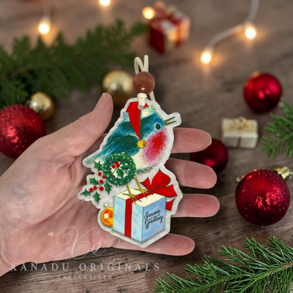 A hand holds a handmade Christmas ornament shaped like a robin bird perched on a gift box, decorated with red ribbon, green holly, and a blue-green bird body. The background shows a rustic wooden surface with red ornaments, green pine branches, and warm string lights.