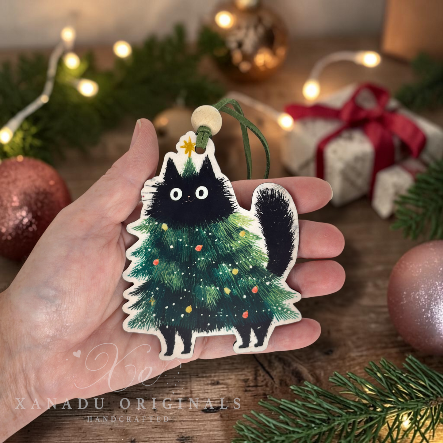 A hand holds a Christmas ornament shaped like a black cat dressed as a green Christmas tree, with small colorful ornaments and a gold star on its head. The background shows a cozy holiday scene with pine branches, warm string lights, gift boxes with red ribbons, and metallic ornaments on a wooden surface.