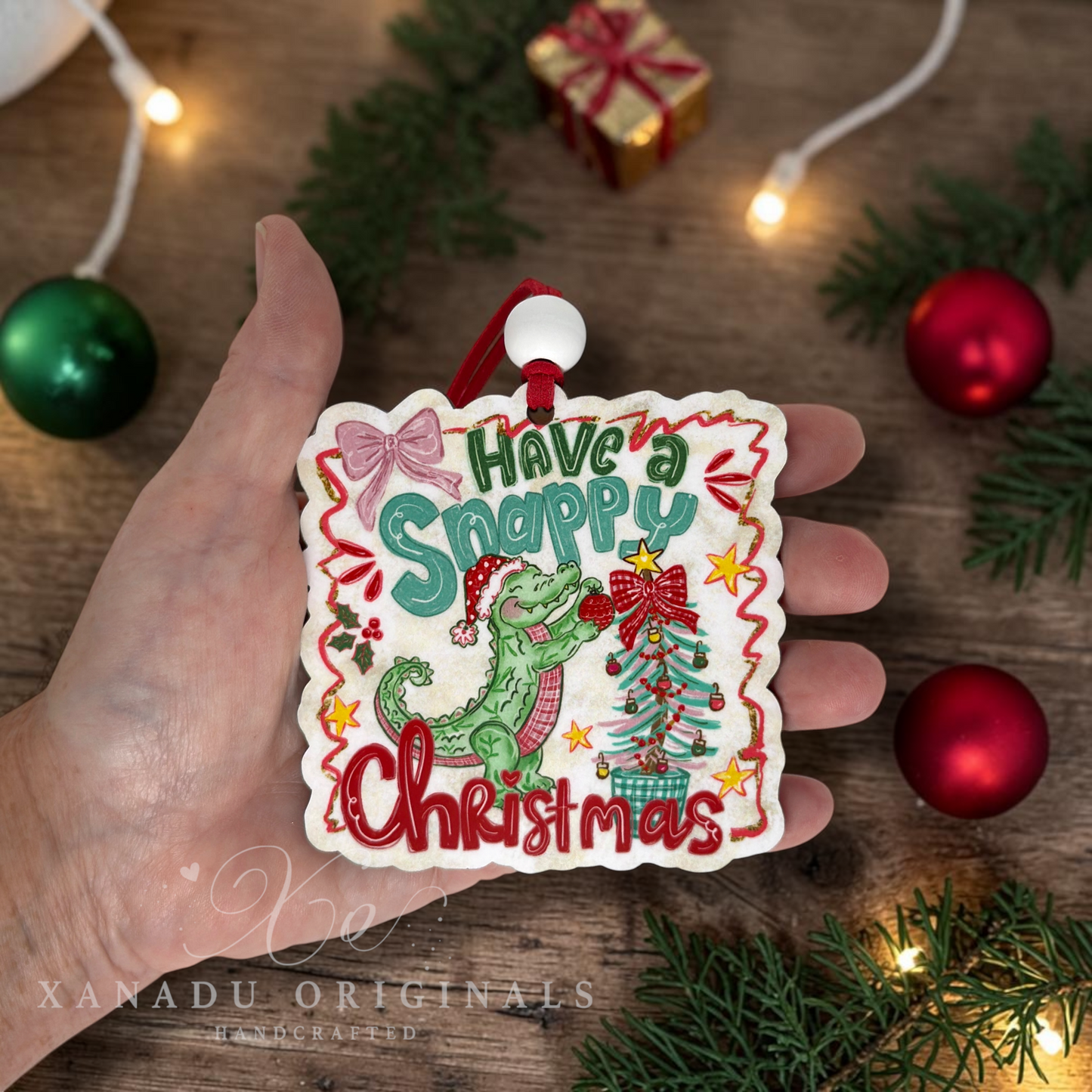 A hand holds a square Christmas ornament with the phrase 'Have a Snappy Christmas' in decorative lettering, featuring a green dinosaur and a small decorated tree. The background shows a wooden surface with holiday lights, pine branches, red and green ornaments, and a wrapped gift box.