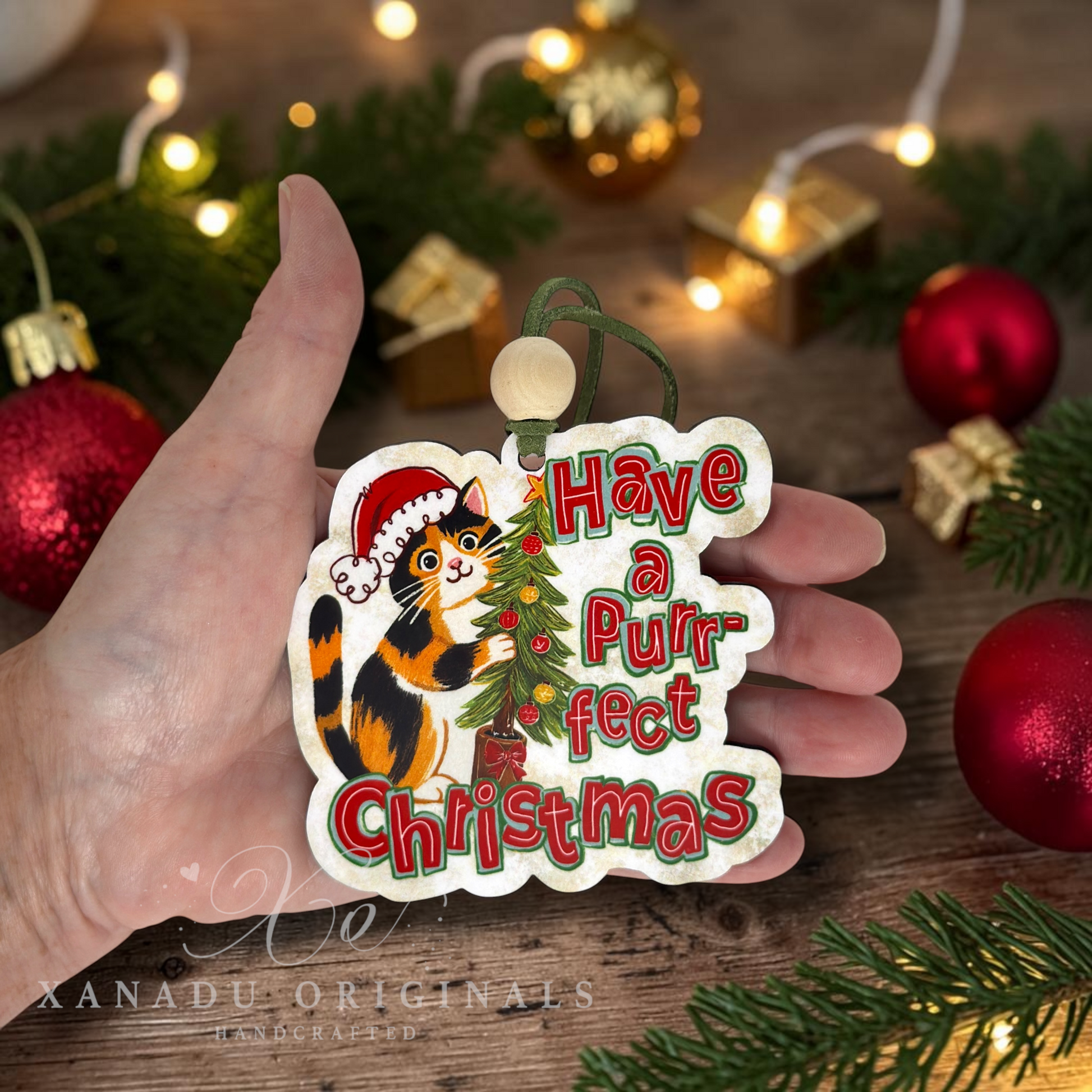 A hand holds a scalloped Christmas ornament shaped like a cat, printed with the phrase 'Have a Purrfect Christmas' in red and green lettering, alongside an illustration of a black, orange, and white cat wearing a Santa hat next to a small decorated tree. The background is a rustic wooden surface with blurred red ornaments, pine branches, and warm string lights.