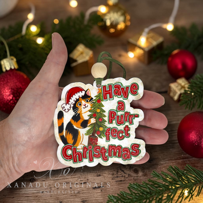 A hand holds a scalloped Christmas ornament shaped like a cat, printed with the phrase 'Have a Purrfect Christmas' in red and green lettering, alongside an illustration of a black, orange, and white cat wearing a Santa hat next to a small decorated tree. The background is a rustic wooden surface with blurred red ornaments, pine branches, and warm string lights.