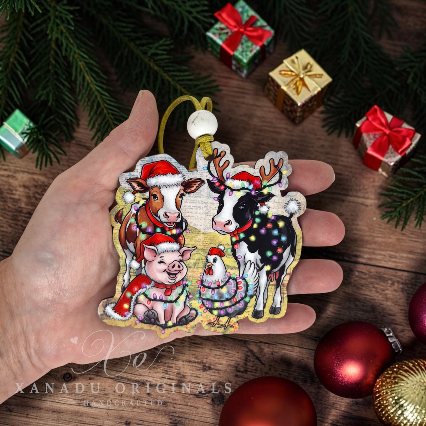 A hand holds a die-cut Christmas ornament featuring a festive group of animals wearing Santa hats and holiday lights. The ornament is surrounded by holiday decorations, including pine branches, wrapped gifts, and red ornaments on a wooden surface.