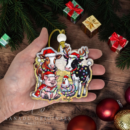 A hand holds a die-cut Christmas ornament featuring a festive group of animals wearing Santa hats and holiday lights. The ornament is surrounded by holiday decorations, including pine branches, wrapped gifts, and red ornaments on a wooden surface.