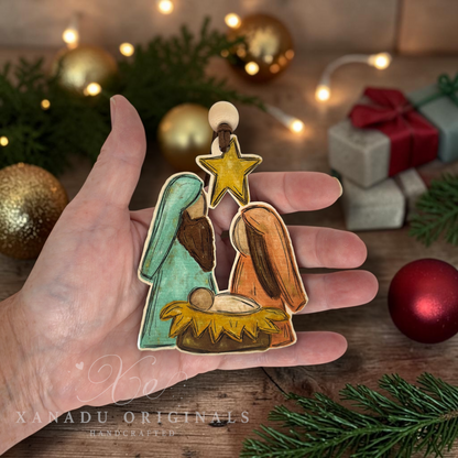 A hand holds a hand-painted wooden nativity ornament featuring Mary, Joseph, baby Jesus, and a gold star. The background is a rustic wooden surface decorated with pine branches, gold Christmas ornaments, and a wrapped gift, with a 'XANADU ORIGINALS' watermark at the bottom.