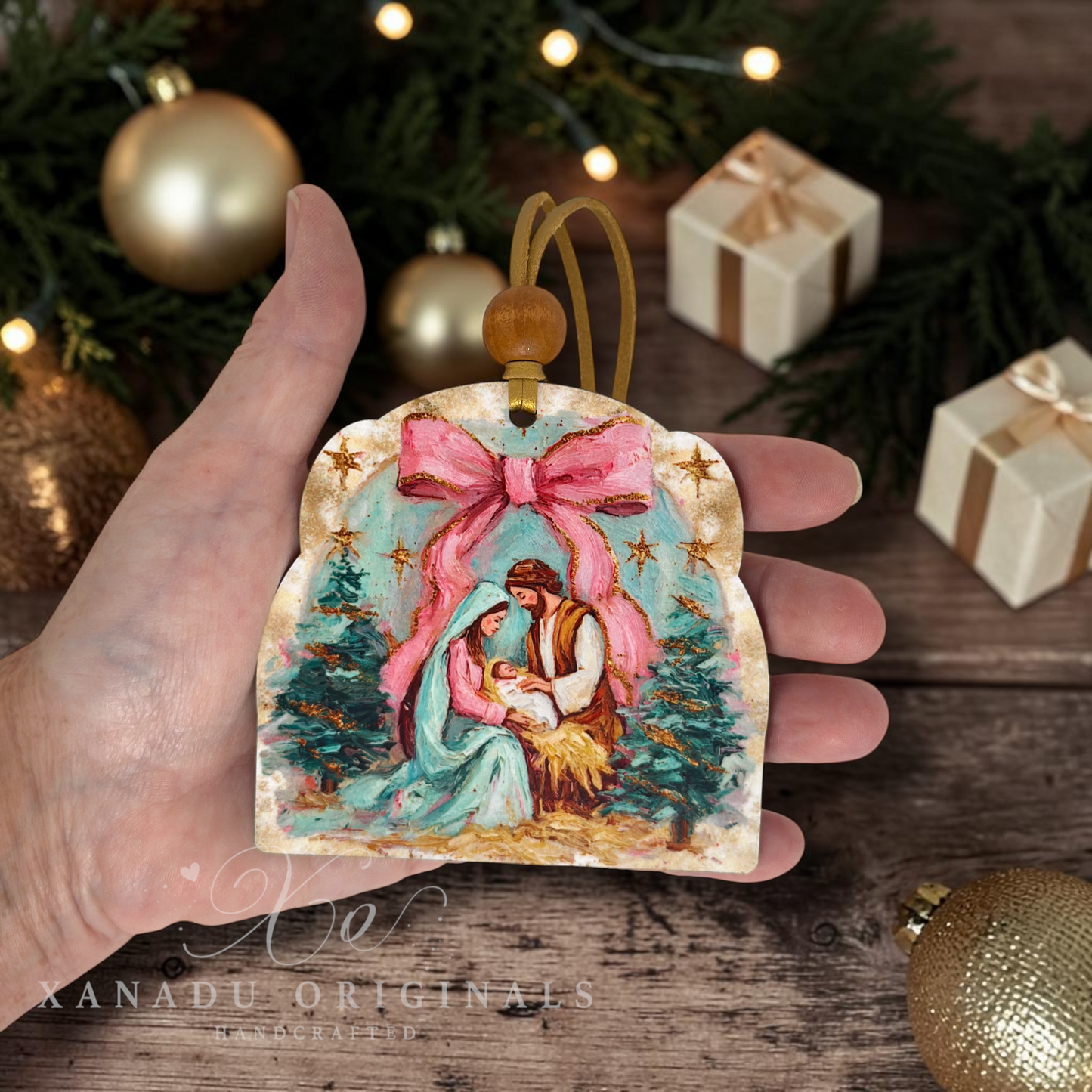 A hand holds a Christmas ornament shaped like a wooden sign, featuring a painted scene of Mary and baby Jesus with a large pink bow, surrounded by festive ornaments and wrapped gifts on a wooden surface.