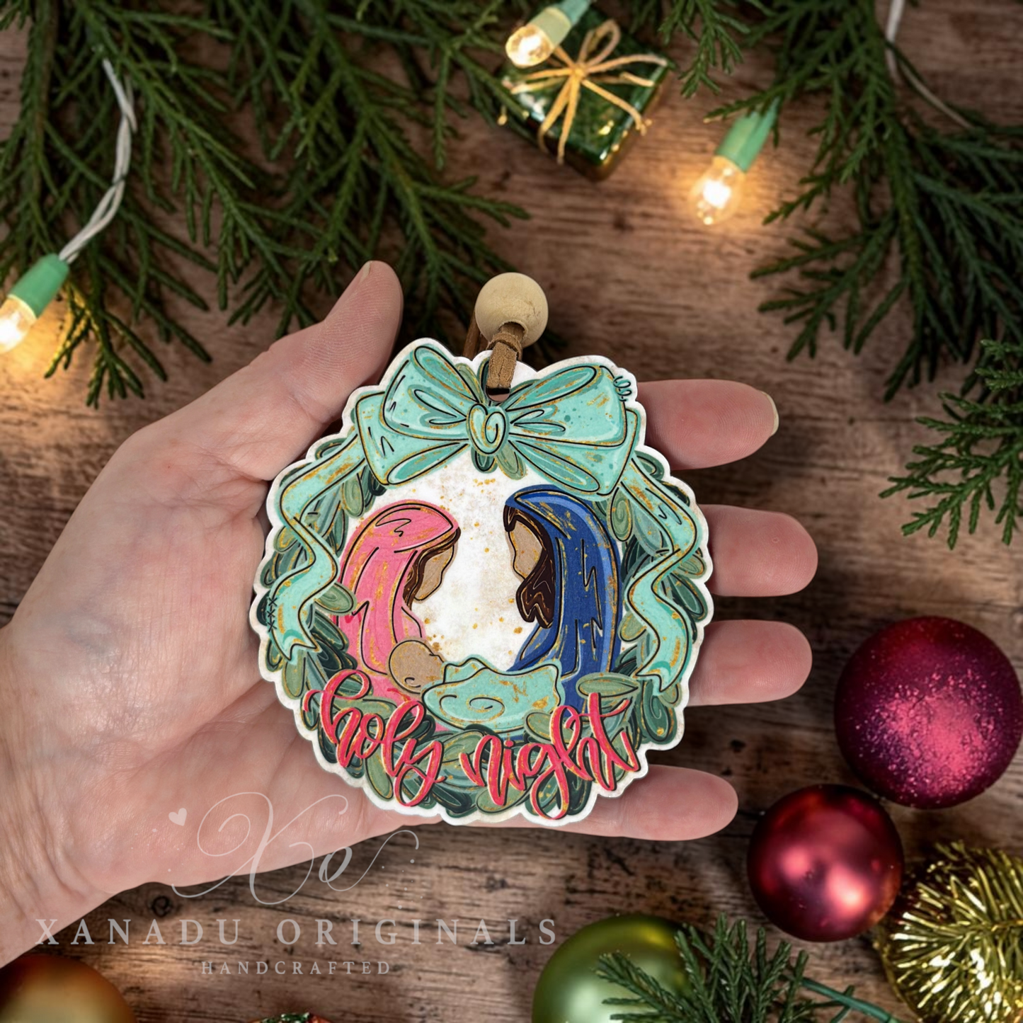 A hand holds a handcrafted Christmas ornament shaped like a wreath, featuring Mary and Joseph in pink and blue robes with the words 'Holy Night' in red. The background is a rustic wooden surface decorated with pine branches, warm string lights, and colorful Christmas ornaments.