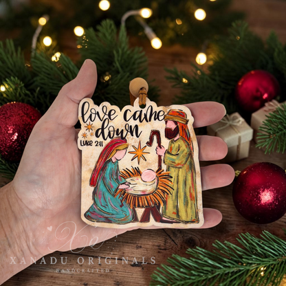 A hand holds a rectangular wooden Christmas ornament featuring a hand-painted nativity scene with the text 'love came down Luke 2:11'. The background is a festive holiday setting with pine branches, red ornaments, and warm string lights.