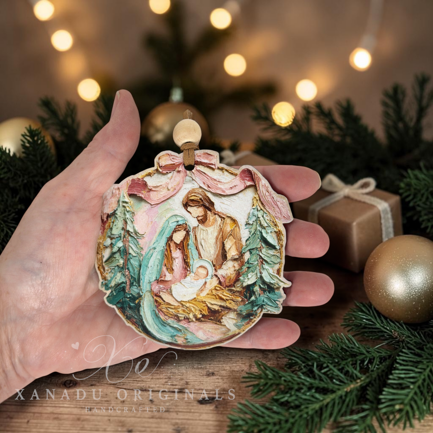 A hand holds a round, hand-painted Christmas ornament depicting the Holy Family (Mary, Joseph, and baby Jesus) in a rustic nativity scene. The ornament features a pink bow at the top and is shown against a festive background with pine branches, gold ornaments, and warm bokeh holiday lights.