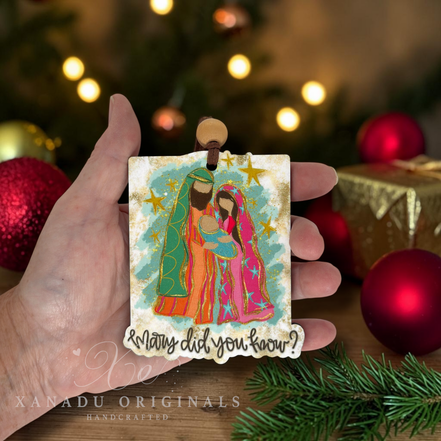 A hand holds a small square Christmas ornament featuring a colorful illustration of Mary, Joseph, and baby Jesus with the text '& Many did you know?'. The background shows a blurred Christmas tree with lights, red ornaments, and a wrapped gift.