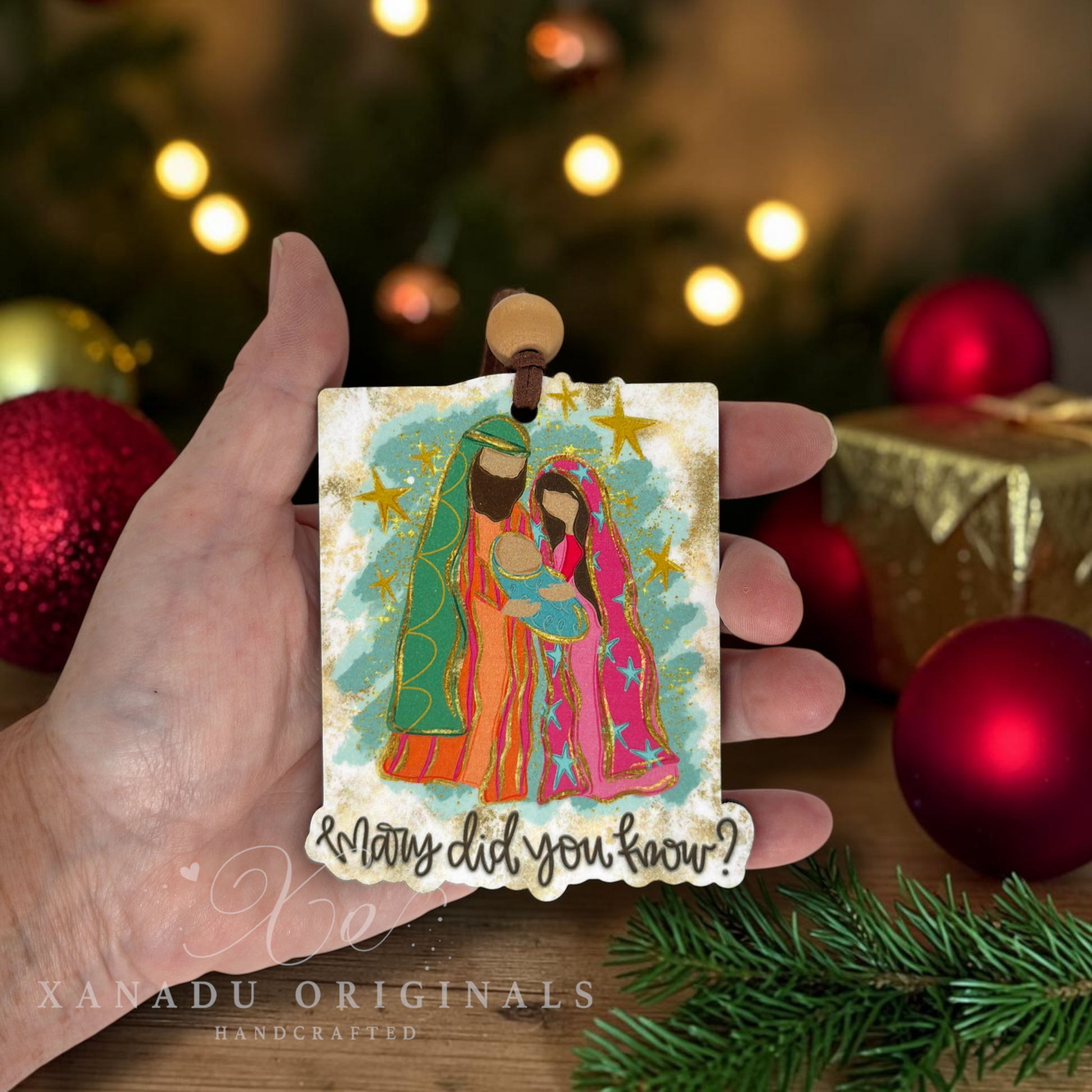 A hand holds a small square Christmas ornament featuring a colorful illustration of Mary, Joseph, and baby Jesus with the text '& Many did you know?'. The background shows a blurred Christmas tree with lights, red ornaments, and a wrapped gift.
